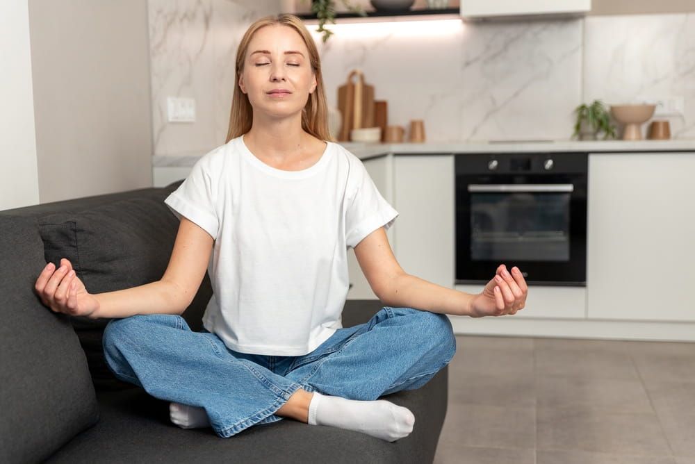 A woman sits on couch, practicing mindfulness with eyes closed. She enjoys a moment of relaxation and meditation at home