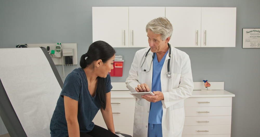 Senior Caucasian male gynecologist in clinic talking to Asian woman give medical history