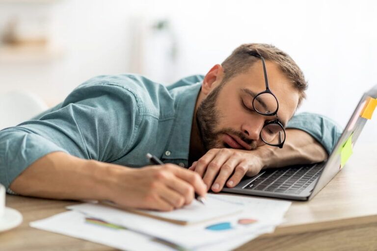 Exhausted millennial man sleeping on his office desk, next to laptop and documents, tired of overworking.